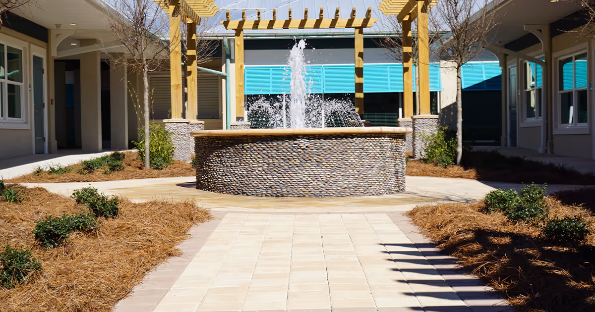 Outdoor courtyard area with a circular stone fountain in the center, surrounded by a paved walkway, small bushes, and trees. The courtyard is framed by a wooden pergola and the exterior walls of the assisted living facility with windows and blue shutters.