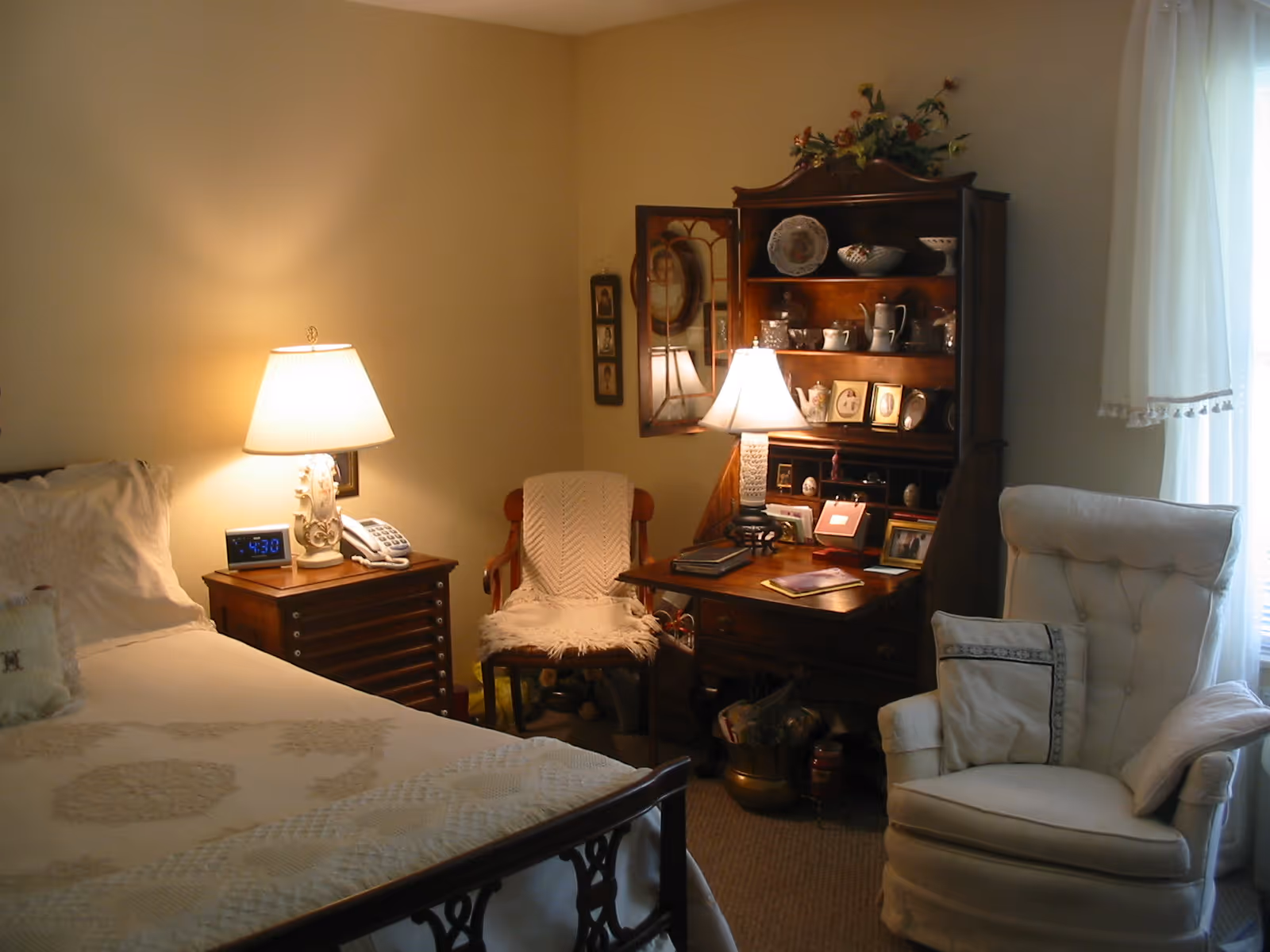 A cozy bedroom with a bed on the left side covered with a white quilt and pillows. Next to the bed is a wooden nightstand with a lamp, a clock showing 4:30, and a telephone. In the corner, there is a wooden chair with a white knitted throw draped over it. To the right, there is a wooden desk with shelves holding decorative plates, teapots, and framed photos. A lamp on the desk illuminates the area. A cushioned armchair with pillows is positioned near a window with sheer white curtains.