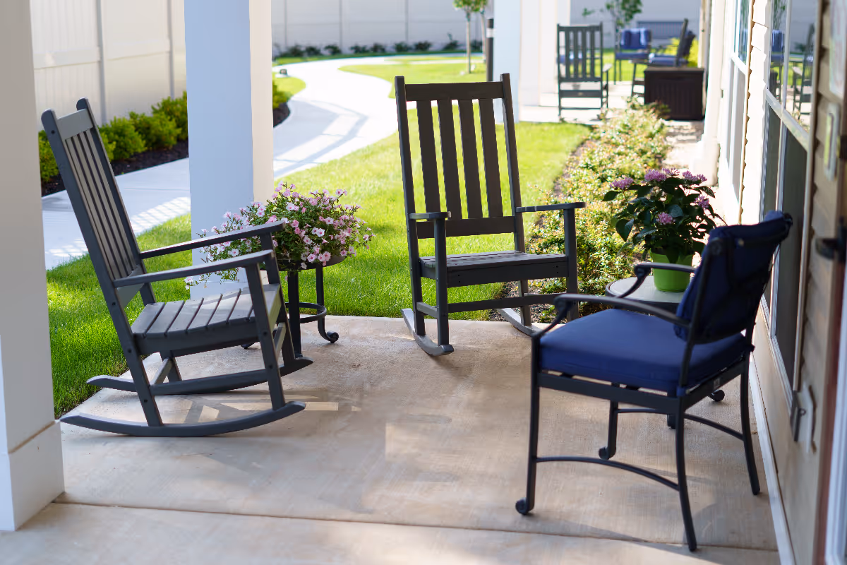 A covered outdoor patio area with three chairs, including two black rocking chairs and one black chair with blue cushions, surrounded by potted flowering plants and greenery. A concrete walkway and well-maintained lawn are visible in the background.