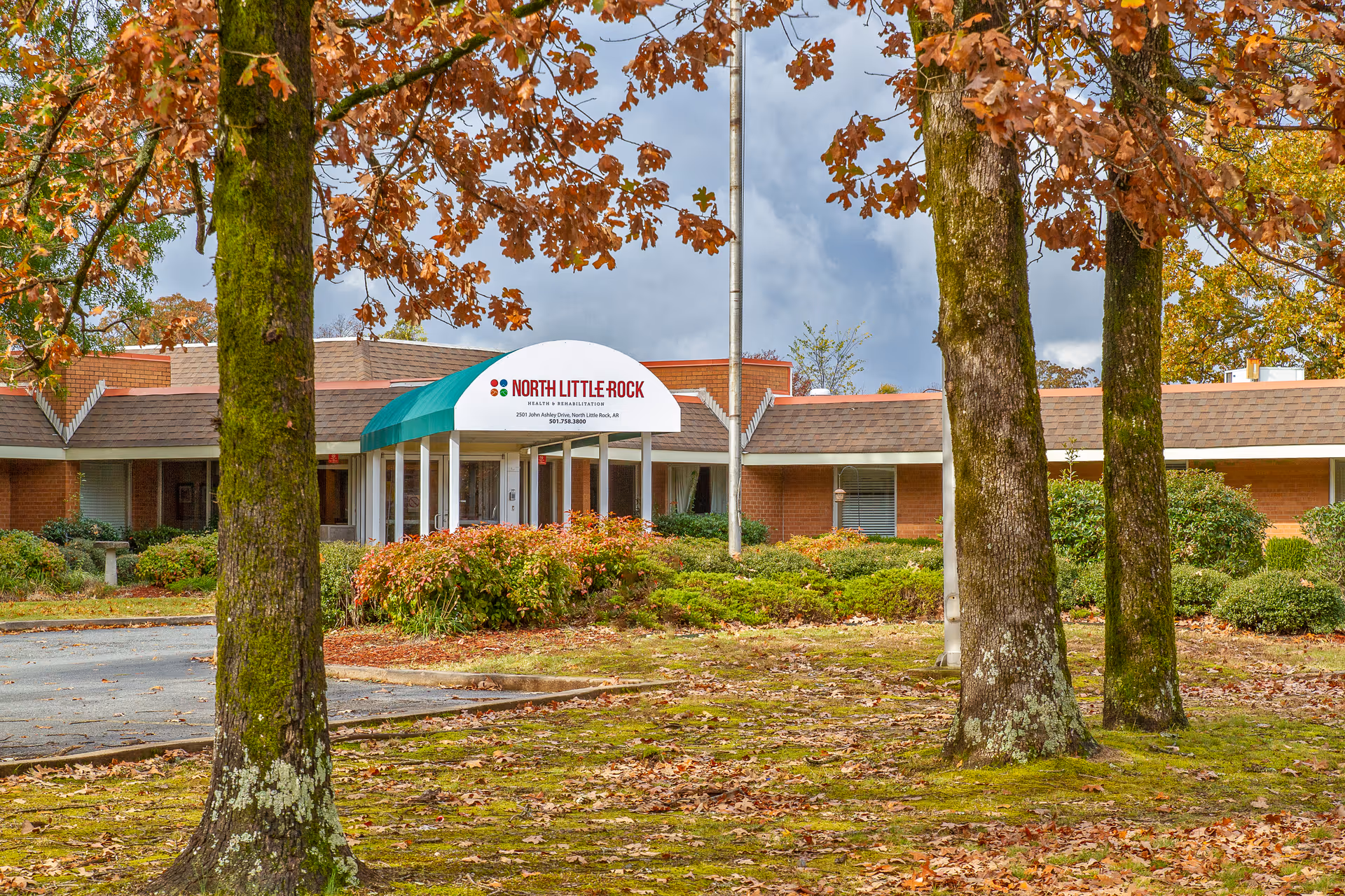 Exterior view of The Blossoms at North Little Rock Rehab & Nursing Center building with a green and white awning over the entrance. The building is surrounded by trees with autumn leaves and landscaped bushes.
