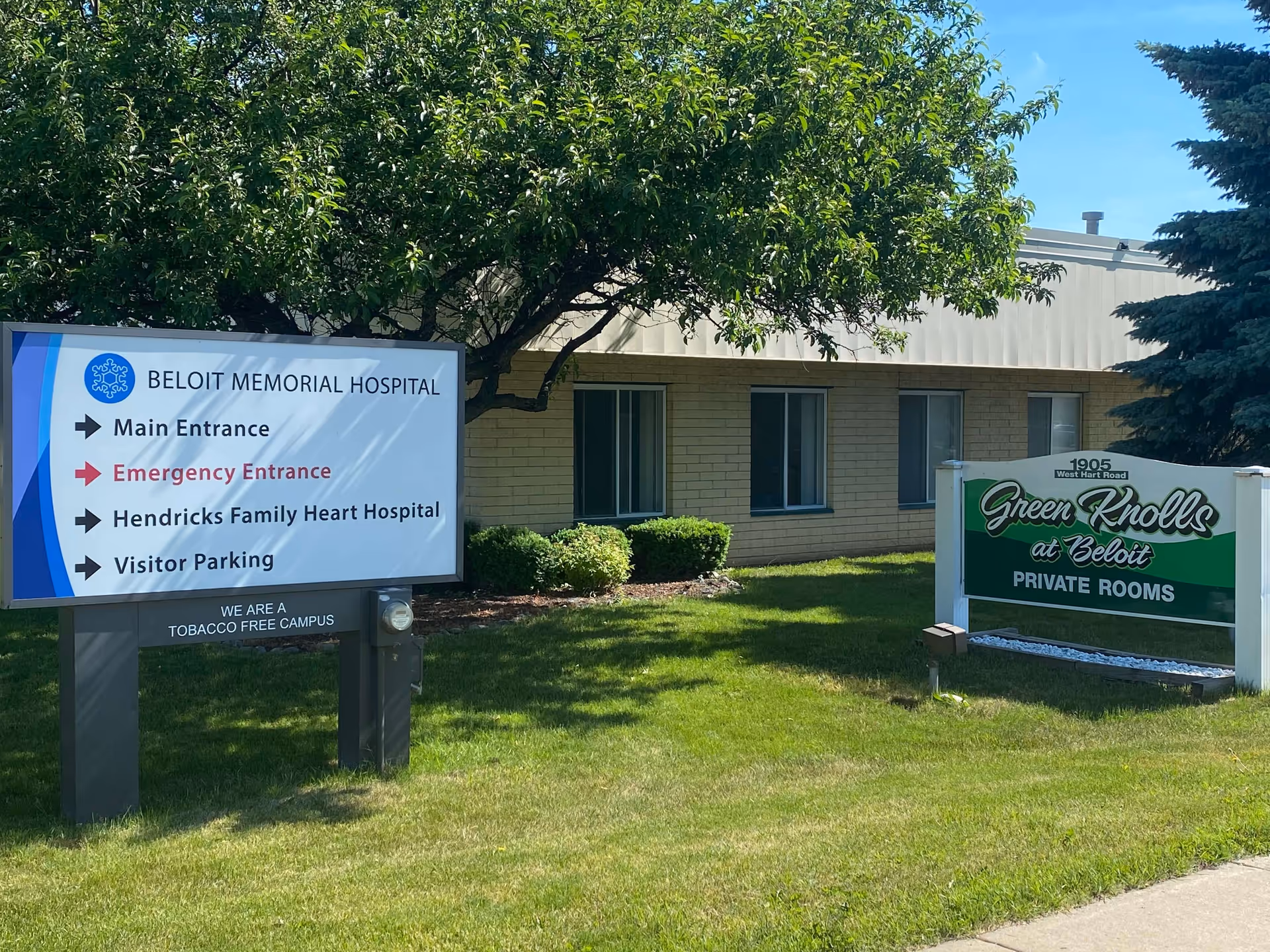 Outdoor view of two signs on a grassy area in front of a building. One sign directs to Beloit Memorial Hospital with arrows for Main Entrance, Emergency Entrance, Hendricks Family Heart Hospital, and Visitor Parking. The other sign reads 'Green Knolls at Beloit PRIVATE ROOMS' with the address 1905 West Hart Road. Trees and bushes are visible around the signs and building.