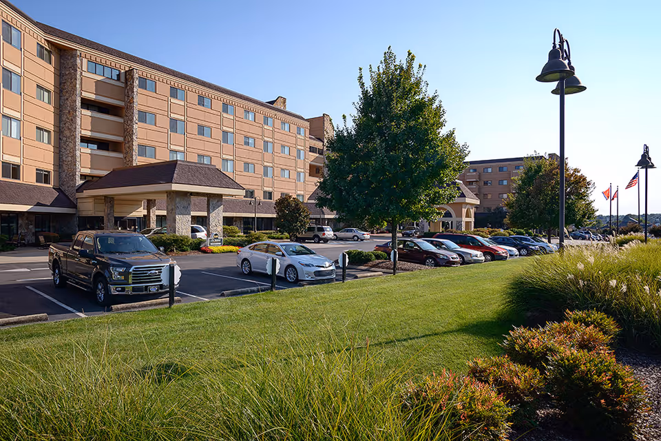 Exterior view of a multi-story senior living facility named Garden Plaza at Cleveland with a parking lot in front, several parked cars, green lawn, trees, and lamp posts under a clear blue sky.