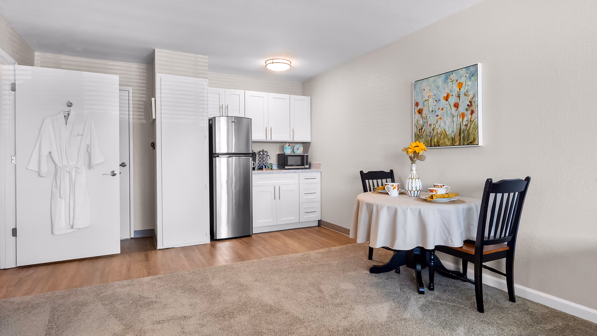A small kitchen and dining area in a senior living facility. The kitchen features white cabinets, a stainless steel refrigerator, a microwave, and a coffee maker. To the right, there is a round dining table covered with a white tablecloth, set with two place settings and a vase of yellow flowers. A colorful floral painting hangs on the wall above the table. A white bathrobe is hanging on the open door to the left.
