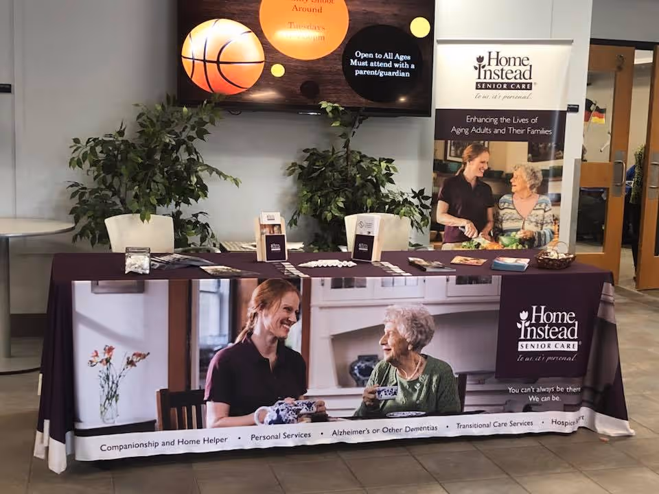 A promotional table setup for Home Instead Senior Care featuring a tablecloth with an image of a caregiver and an elderly woman smiling and drinking tea together. The table has brochures, business cards, and informational materials. Behind the table is a banner with the Home Instead logo and the slogan 'Enhancing the Lives of Aging Adults and Their Families.' There are two potted plants on either side of the table, and a television screen above displaying a basketball graphic and event information.