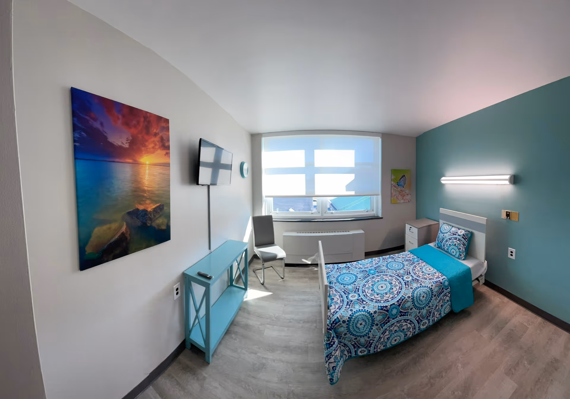 A bright and clean nursing facility bedroom with a single bed covered in a blue patterned bedspread and matching pillow. The room has a teal accent wall with a horizontal light fixture above the bed. There is a window with a white blind letting in natural light, a small nightstand, a chair, a wall-mounted TV, and a blue console table. A colorful sunset beach painting hangs on the wall opposite the bed.