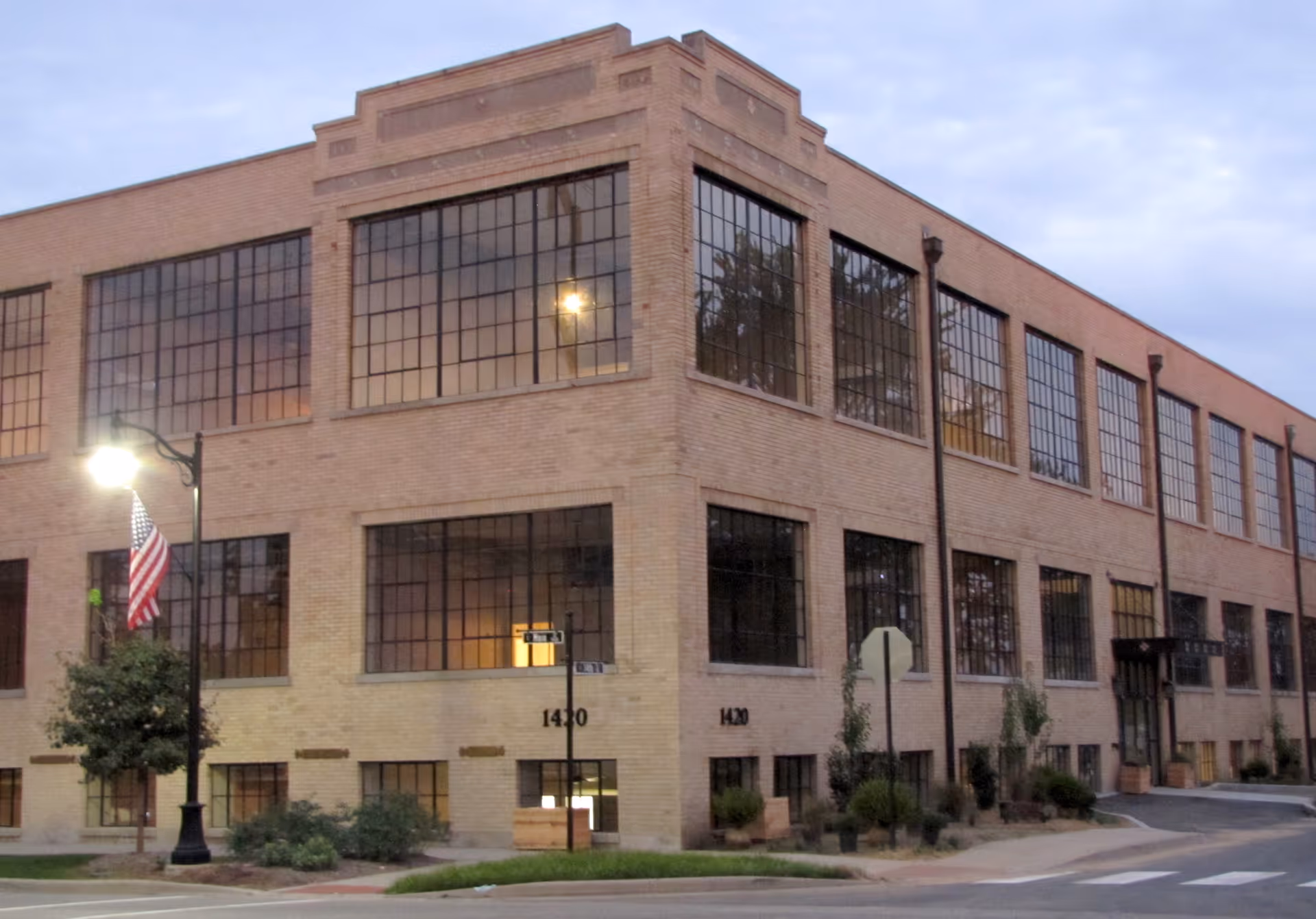 Exterior view of a large two-story brick building with many large windows, a street lamp with an American flag, and some greenery along the sidewalk. The building has the address number 1420 visible on the corner.
