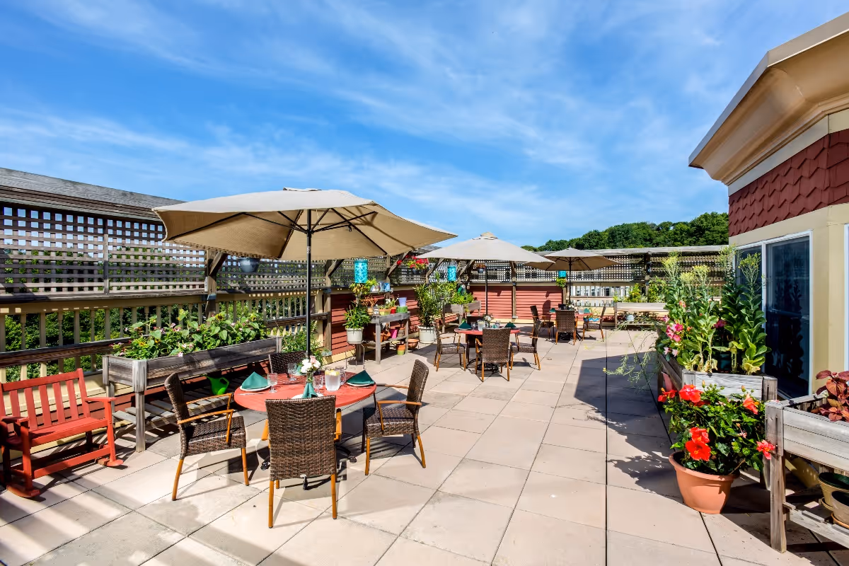 Outdoor patio area with several tables and chairs under large beige umbrellas, surrounded by potted plants and flowers, with a wooden lattice fence and a clear blue sky in the background.
