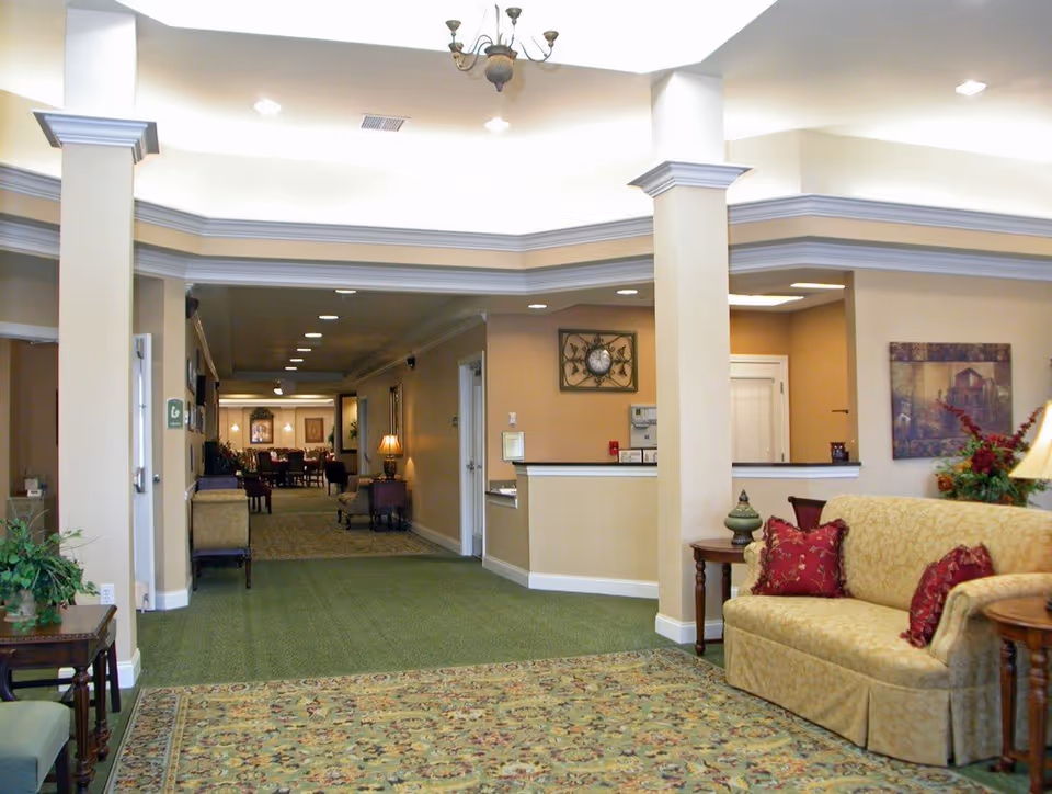 Interior view of a senior living facility lobby area with beige walls, green carpet, a patterned area rug, and a yellow sofa with red pillows. There are side tables with lamps and decorative items, a chandelier on the ceiling, and a hallway leading to a dining area with tables and chairs in the background.