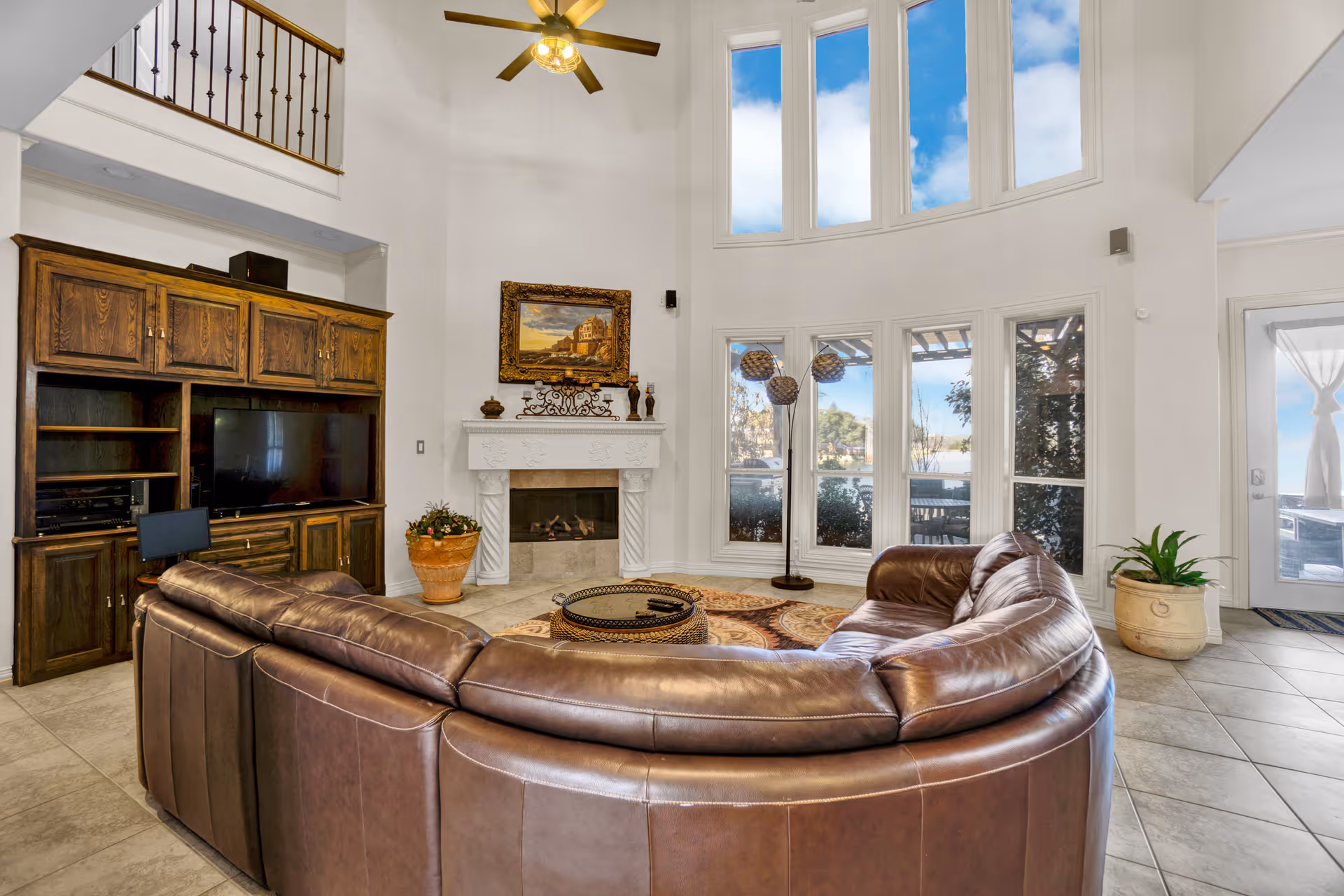 Spacious living room with a curved brown leather sectional facing a TV cabinet and white fireplace beneath tall arched windows.