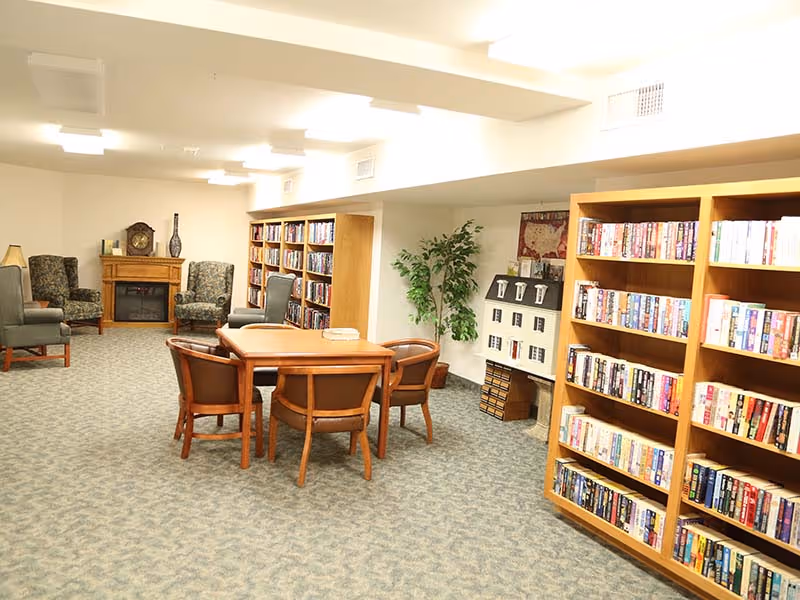 A cozy senior living community library and lounge area with bookshelves filled with books, a wooden table surrounded by six chairs, two armchairs near a fireplace with a clock and decorative vase on the mantel, and a potted plant next to a dollhouse on a small table.