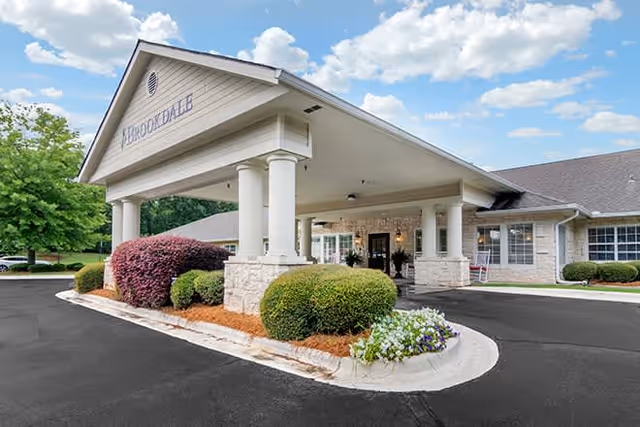 Exterior view of the entrance to Brookdale Columbus senior living facility featuring a covered drop-off area with white columns, landscaped bushes, and a clear blue sky with some clouds.