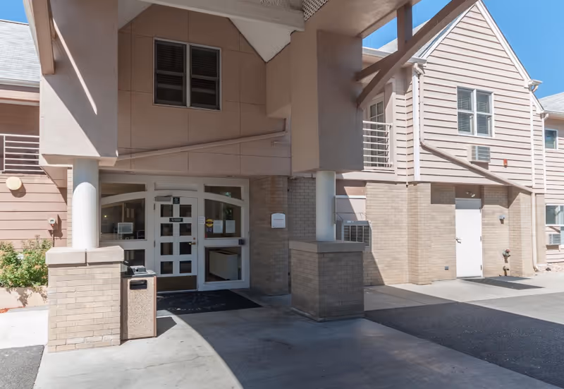 Entrance to a senior living facility with a covered drop-off area supported by columns. The building exterior features beige brick and light pink siding with multiple windows. There is a trash bin near the entrance and a sign on the wall next to the door.