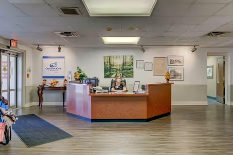Reception area of Christian Health Center Louisville with a woman wearing a mask sitting behind a wooden front desk. The room has light-colored walls, wood flooring, and ceiling lights. There is a sign on the left wall that reads 'Christian Care Communities' and framed pictures on the wall behind the desk. A small table with decorations is visible near the entrance door on the left.