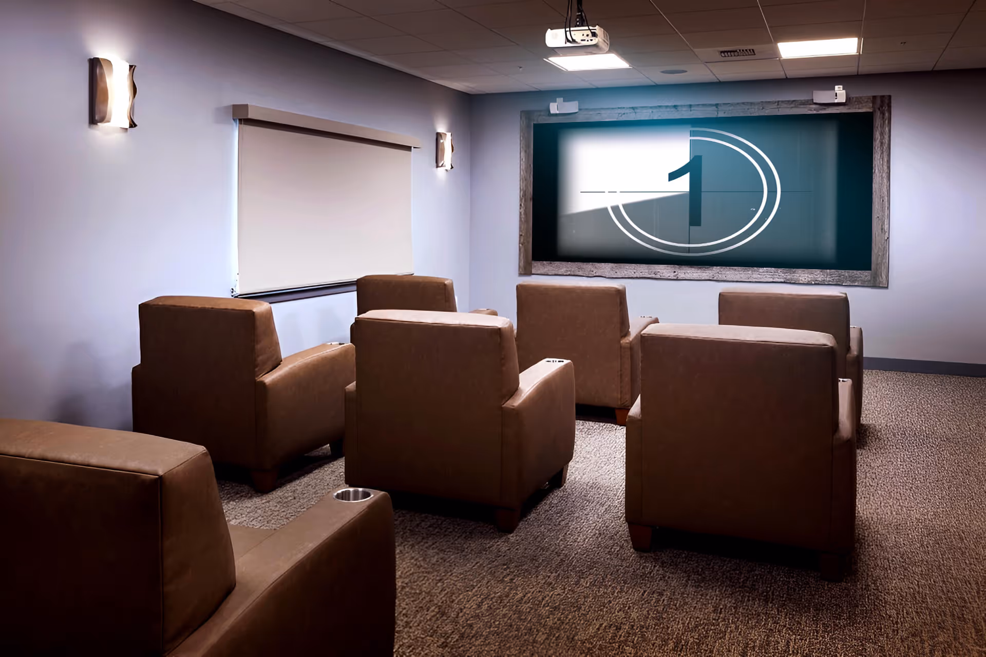 A small theater room with six brown leather armchairs arranged in two rows facing a large screen showing a countdown number 1. The room has light-colored walls, carpeted floor, two wall-mounted lights, and a ceiling-mounted projector.