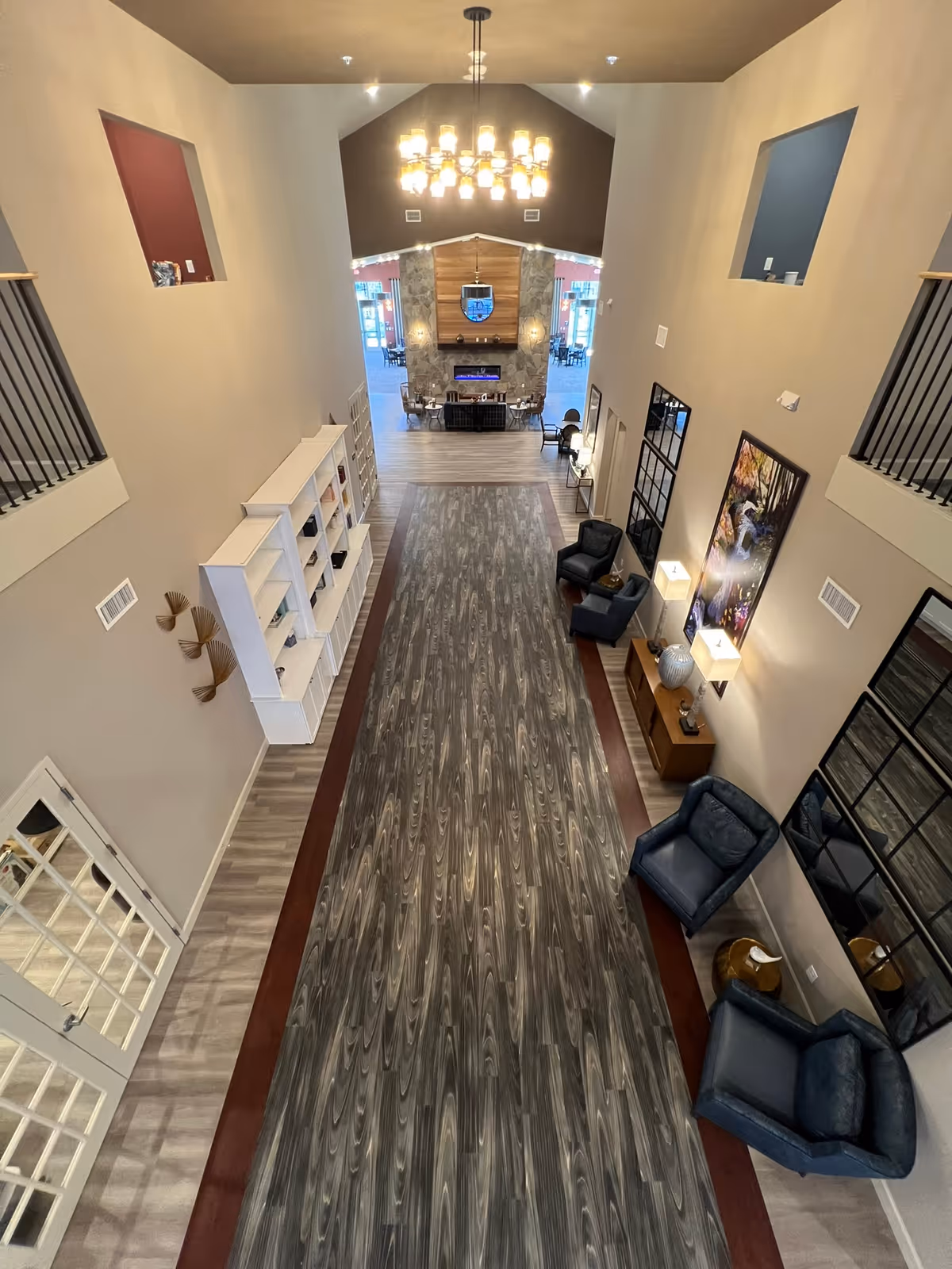 View down a tall open interior atrium with wood-patterned flooring, seating areas, bookshelves, a chandelier, and a fireplace at the far end.