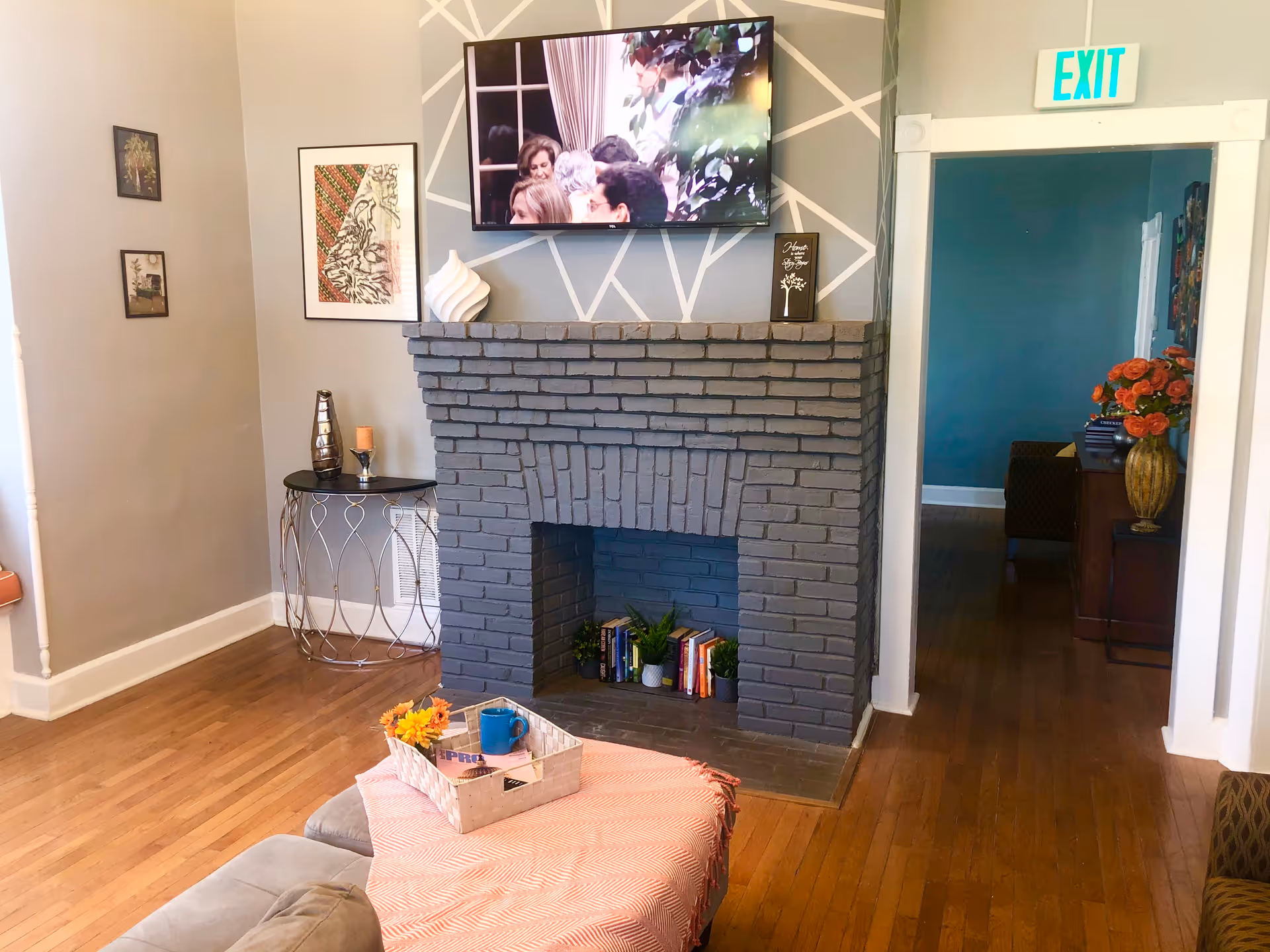 A cozy living room area with a gray brick fireplace, a TV mounted above it displaying a group of people, and decorative items on the mantel. There is a small round table with a candle and vase to the left of the fireplace. A tray with a blue mug and flowers sits on a pink cloth on a cushioned ottoman in the foreground. The room has wooden floors and light-colored walls with framed artwork. An open doorway leads to another room with a blue wall and a vase of orange flowers on a table.