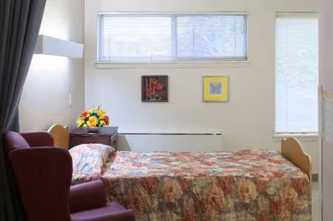 Bedroom in care facility with floral bedspread, burgundy chair, and flower arrangement.