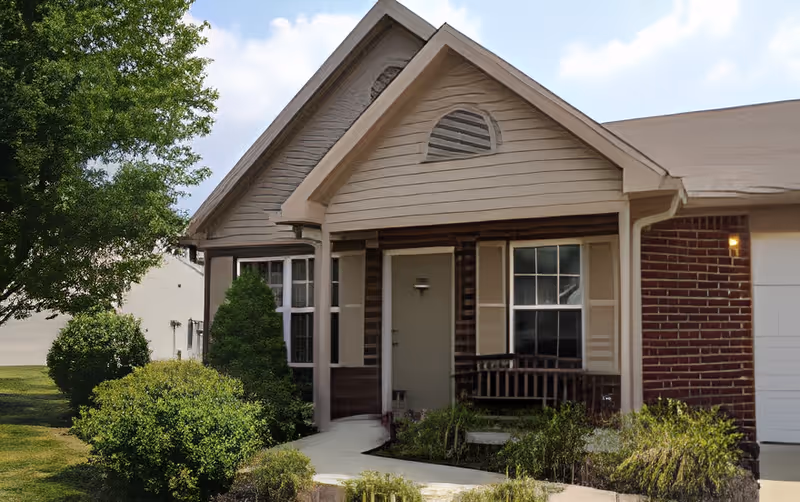Exterior view of a single-story residential building with a small covered porch, a front door, windows with shutters, and surrounding greenery including bushes and a tree under a partly cloudy sky.