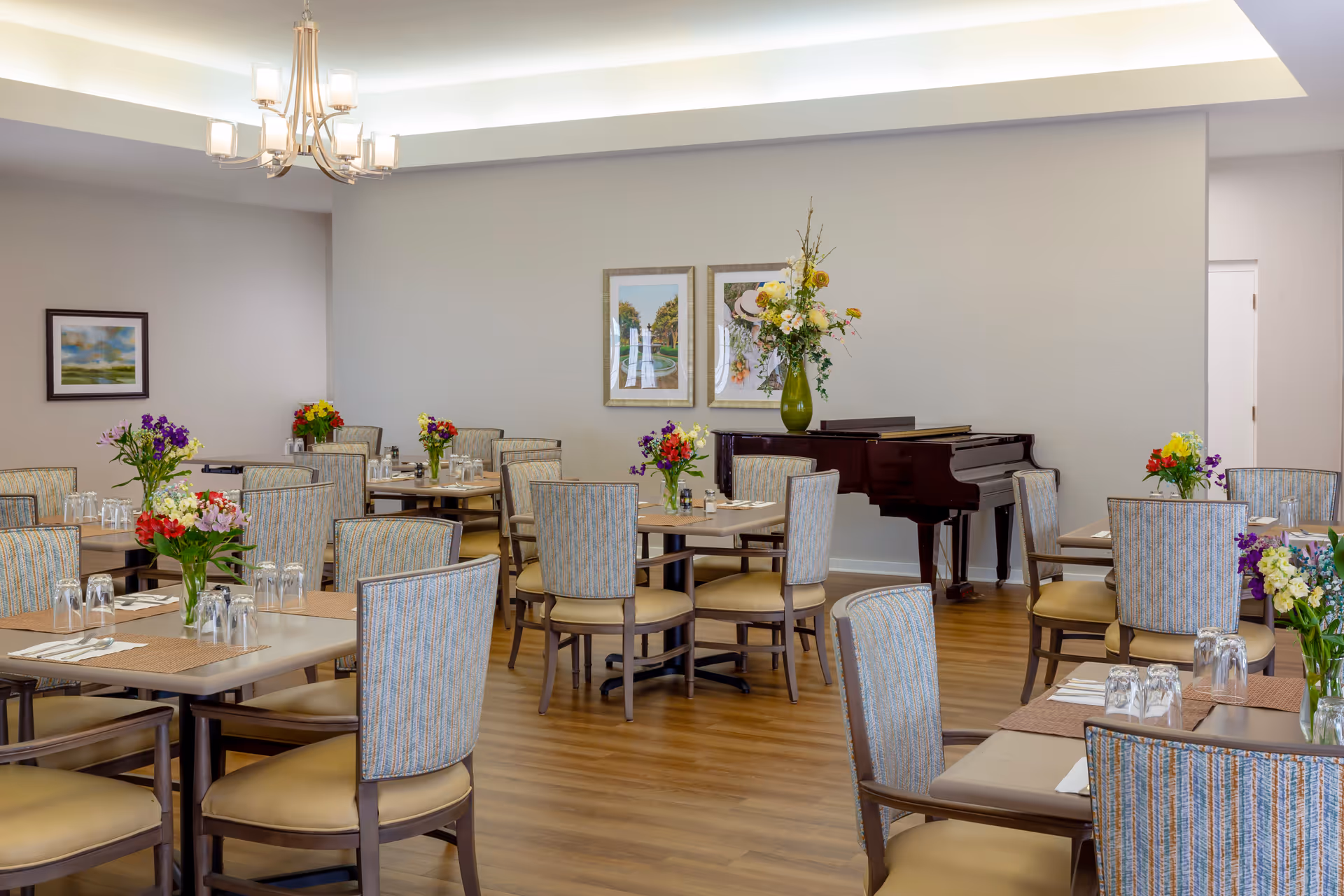 Bright dining room with multiple tables and chairs, floral centerpieces, and a grand piano against the back wall.