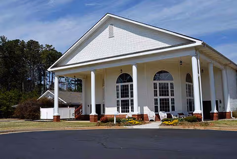 Exterior view of a white building with large columns and arched windows, surrounded by a paved driveway and some landscaping with flowers and bushes under a blue sky.