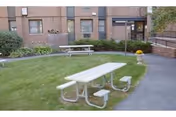 Grassy courtyard with picnic tables and attached benches outside a brick senior living building entrance.