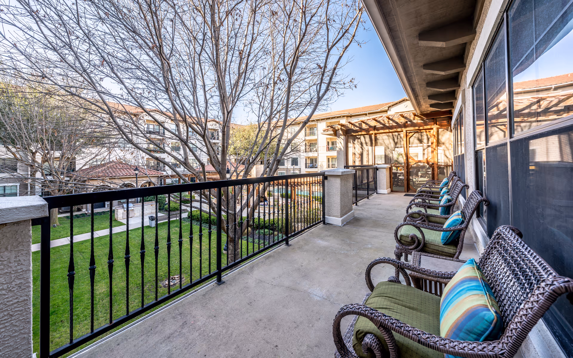 Outdoor balcony area with several wicker chairs featuring green cushions and colorful striped pillows. The balcony overlooks a landscaped courtyard with green grass, trees, and a walking path. The building has a beige exterior with large windows and a pergola-covered entrance at the far end of the balcony.