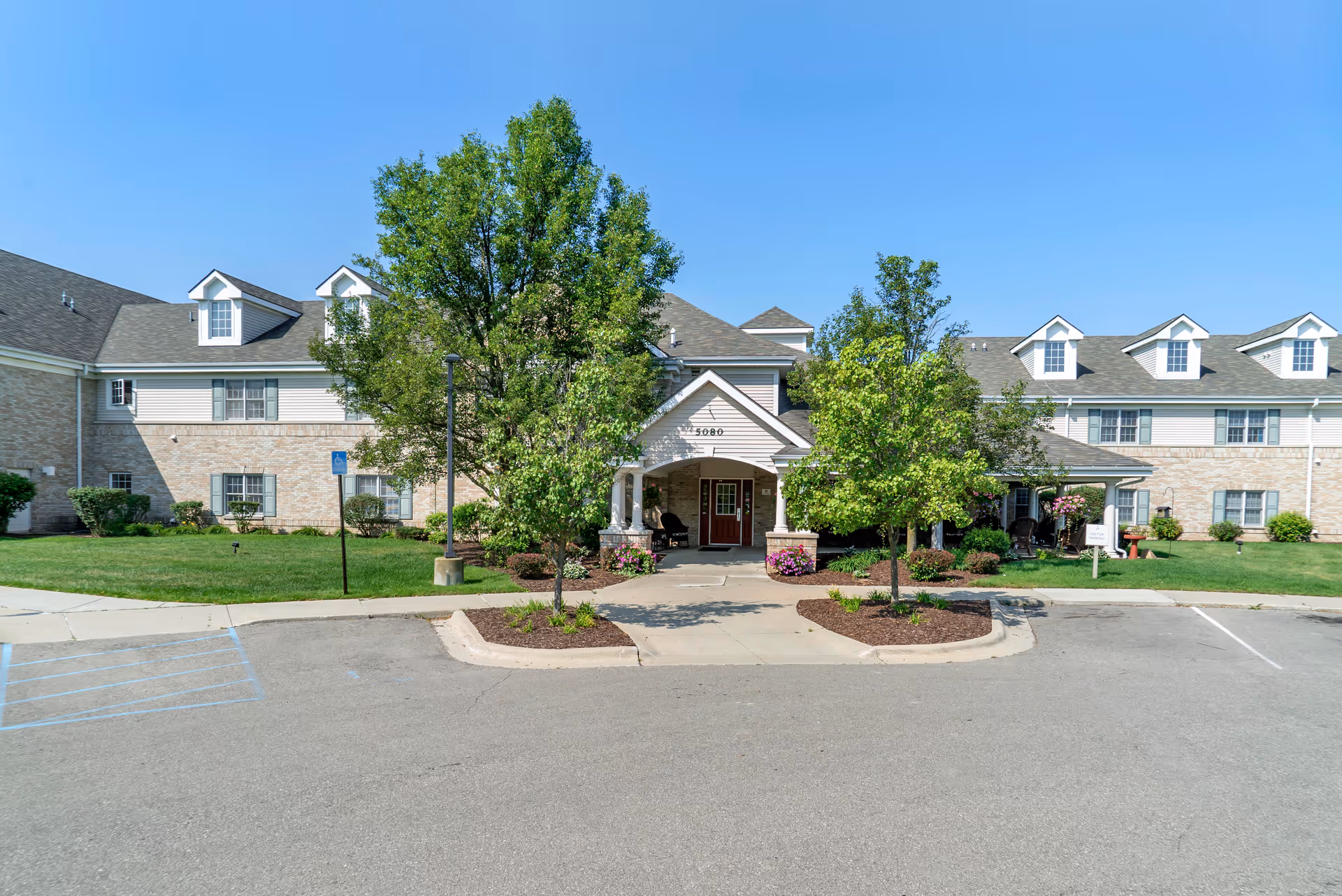 Front exterior view of Brookdale Grand Blanc facility with a paved driveway, landscaped greenery including trees and bushes, and a clear blue sky.