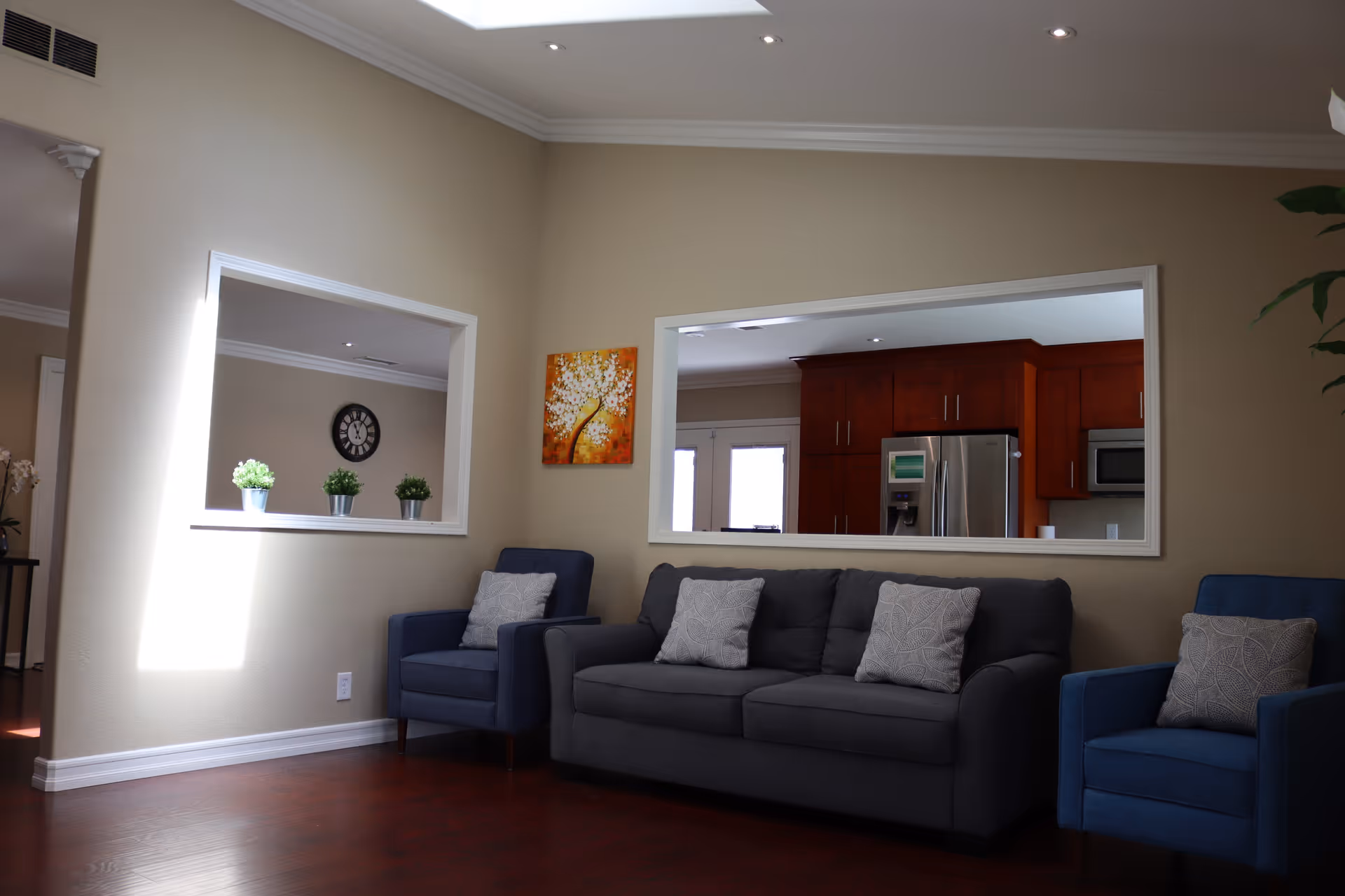 A cozy living room with a dark gray sofa and two blue armchairs, each with patterned cushions. The walls are beige, and there are two large rectangular windows opening into adjacent rooms. One window shows a kitchen with wooden cabinets and a stainless steel refrigerator, while the other window reveals a room with a clock on the wall and three small potted plants on the windowsill. A colorful painting of a tree with white blossoms hangs on the wall between the windows. The floor is dark wood, and the ceiling has recessed lighting.