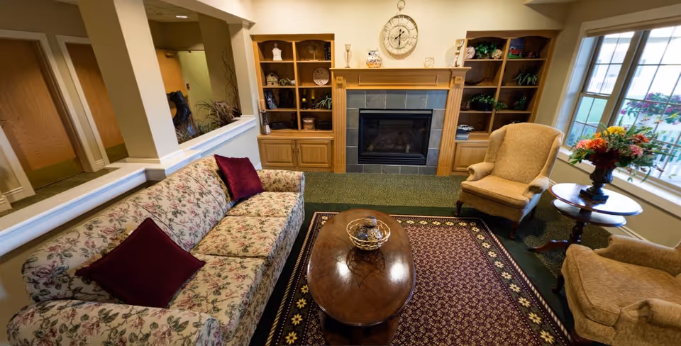 A cozy living room area with a floral patterned sofa adorned with two burgundy pillows, two beige armchairs, a wooden coffee table on a patterned rug, built-in wooden shelves flanking a fireplace with a clock above it, and a window with a flower arrangement on a side table.