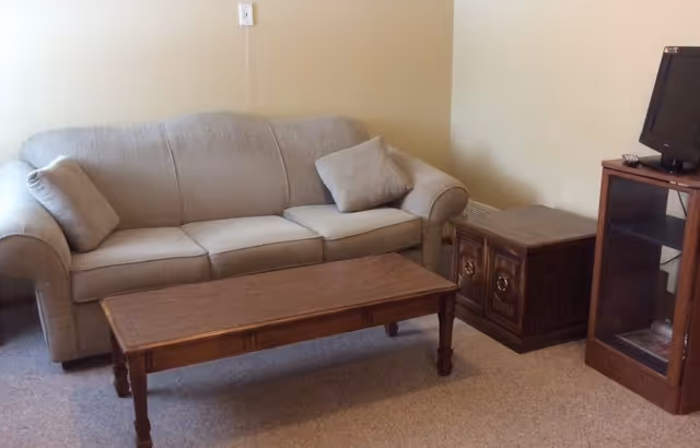 A small living room with a beige couch with two cushions, a wooden coffee table in front of it, a wooden side table next to the couch, and a wooden TV stand with a small television on top. The walls are plain and light-colored, and the floor is carpeted.