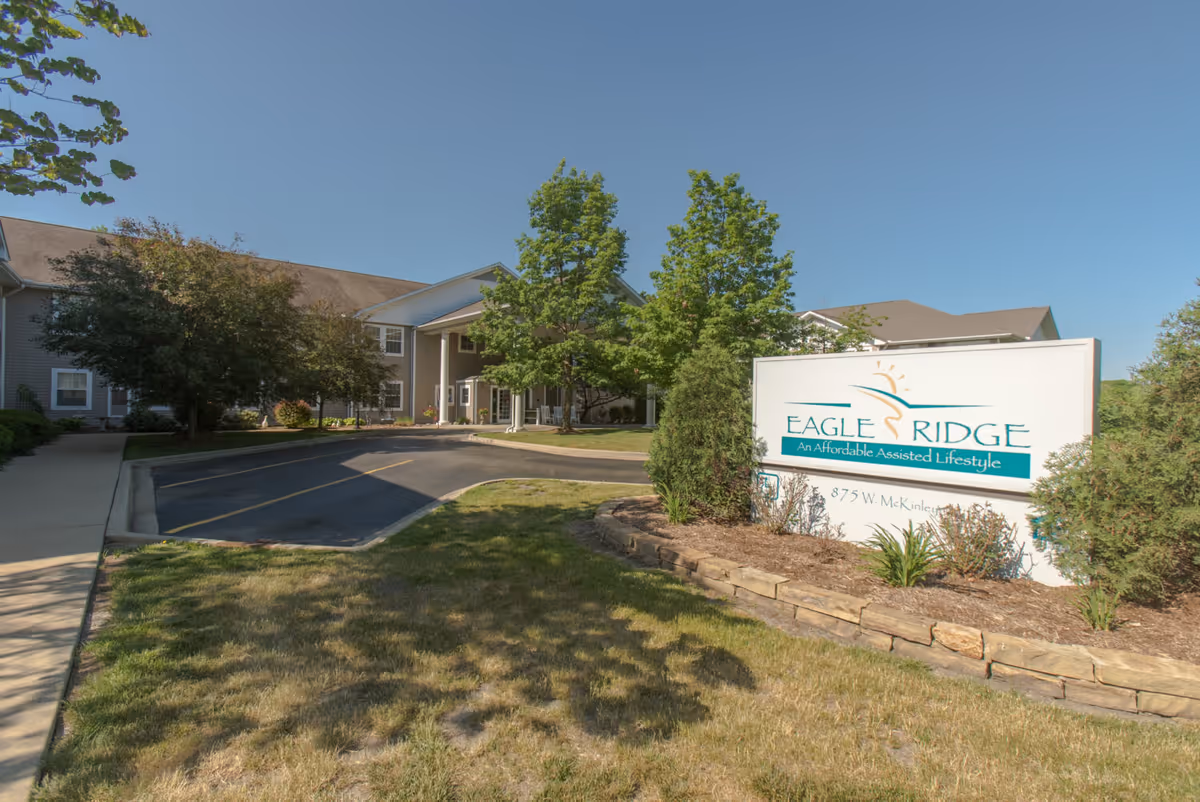 Exterior view of Eagle Ridge of Decatur, an assisted living facility, showing the building entrance with a covered driveway, surrounded by trees and landscaping under a clear blue sky. A large sign in the foreground displays the facility name and slogan.