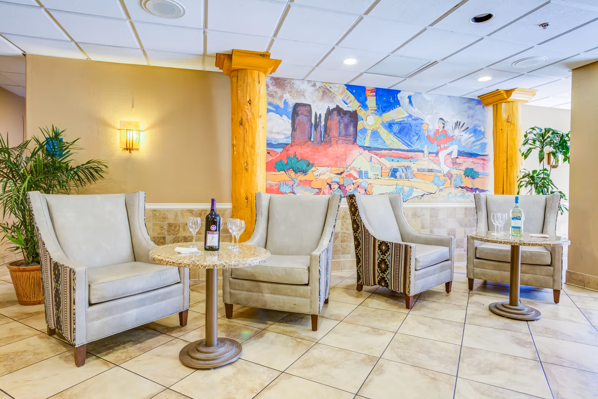 A cozy seating area with four beige armchairs arranged around two small round tables. Each table has a bottle of wine and wine glasses. The room features tiled flooring, wooden columns, potted plants, and a colorful mural on the wall depicting a southwestern desert scene with rock formations, a sun, and a dancing figure.