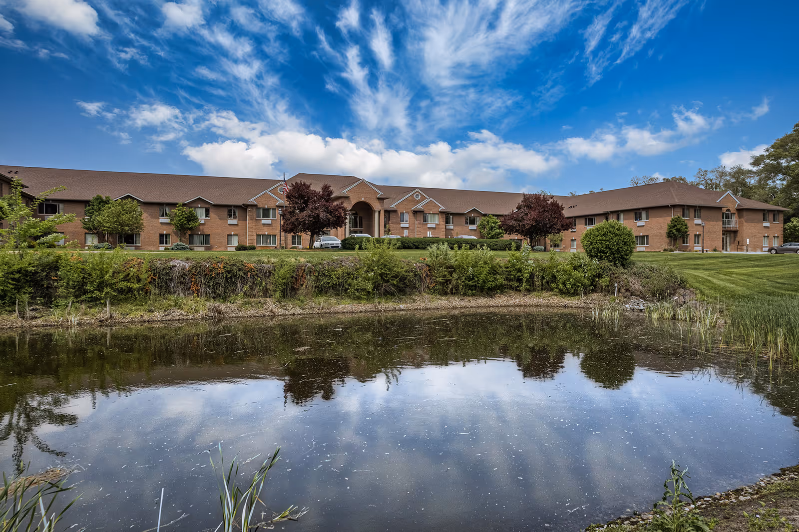 A large two-story brick building with a brown roof, surrounded by green trees and bushes, reflected in a pond in the foreground under a blue sky with scattered clouds.
