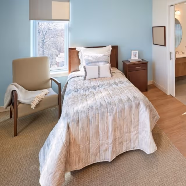 A single bed with a wooden headboard is made with a patterned bedspread and three pillows. Next to the bed is a wooden nightstand with a framed photo and a small item on top. To the left of the bed is a beige armchair with a light-colored throw blanket draped over one arm. A window with a roller shade is behind the chair, letting in natural light. The walls are painted light blue, and a doorway on the right leads to a bathroom with a sink and mirror visible.