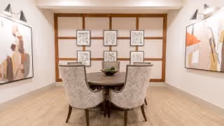 A modern dining room with a round wooden table surrounded by six upholstered chairs. The walls are decorated with abstract art paintings on either side and a grid of framed sketches on the far wall. The floor is light wood, and the room is softly lit with wall sconces.
