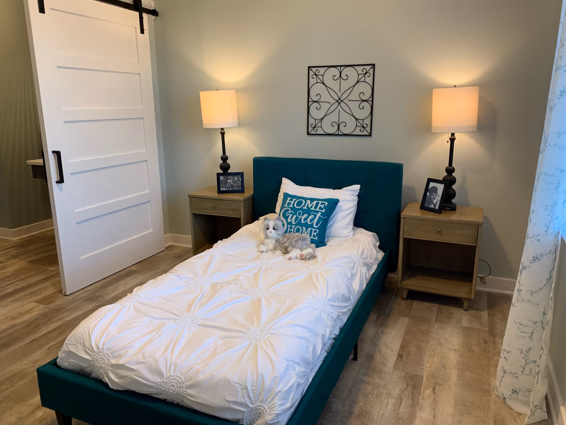 A cozy bedroom with a teal upholstered bed featuring a white comforter and a decorative pillow that says 'Home Sweet Home'. There are two wooden nightstands on either side of the bed, each with a lamp and a framed photo. A decorative metal wall art piece hangs above the bed. The room has light-colored walls and wood flooring, with a sliding white door partially open to an adjacent room.