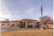 Front exterior of a single-story senior care building with a covered entrance, flagpole, trimmed shrubs, and a circular driveway.