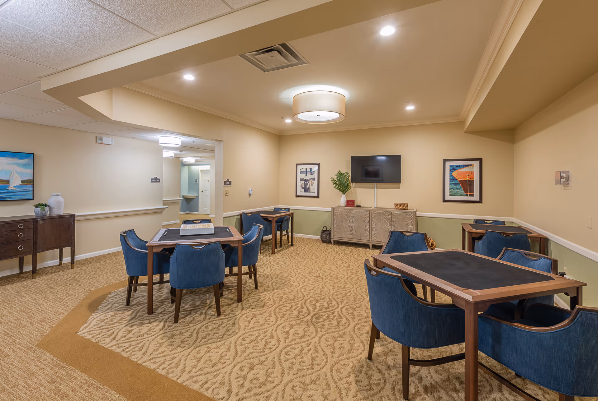 A well-lit common area with multiple tables and blue upholstered chairs arranged for group seating. The room has beige walls with white trim, patterned carpet, framed artwork on the walls, a flat-screen TV mounted above a cabinet, and decorative plants. The space appears clean and inviting, suitable for social activities or gatherings.