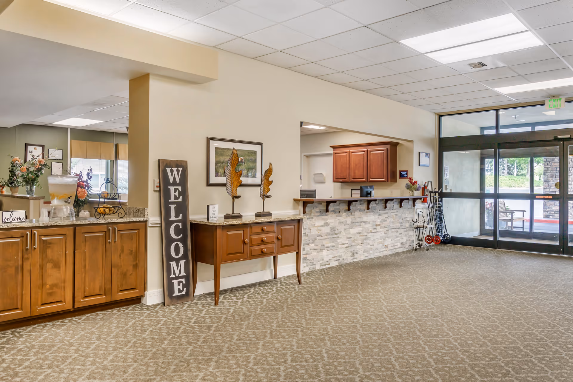 Interior view of a senior living facility lobby area with a reception desk made of stone and wood, a wooden side table with decorative items, a large vertical welcome sign, and a cabinet with a water dispenser and flowers. Large glass doors lead outside, allowing natural light to enter.