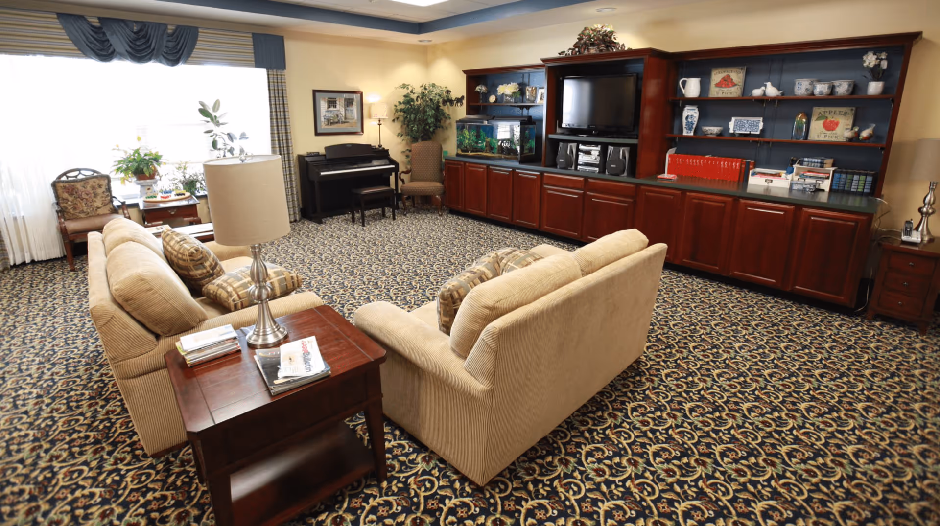 Communal living room with beige sofas facing a TV in built-in wooden cabinetry, a piano by the window, and patterned carpet.