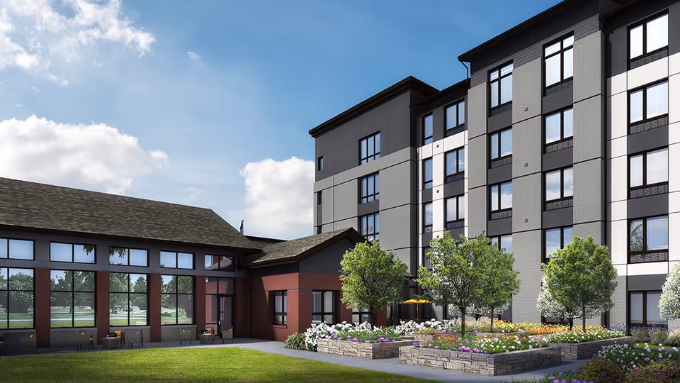 Exterior view of a modern senior living facility with a multi-story building and a single-story section with large windows. The foreground features a well-maintained garden with flower beds, small trees, and a grassy lawn under a partly cloudy sky.