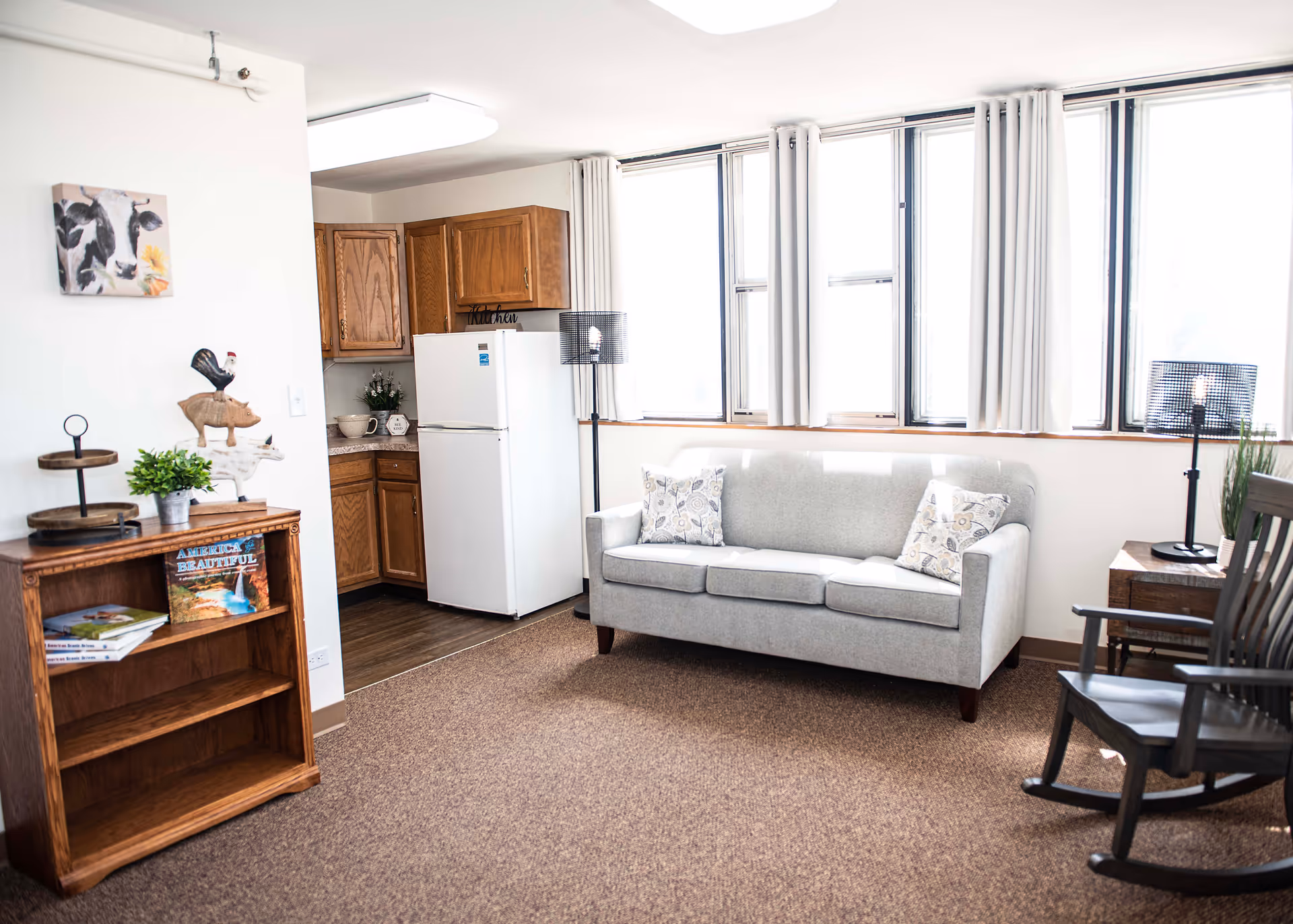 A bright living area with a light gray sofa adorned with two patterned pillows, a wooden rocking chair, and a small wooden side table with a lamp. The room has large windows with white curtains letting in natural light. To the left, there is a small kitchen area with wooden cabinets and a white refrigerator. A wooden bookshelf with books and decorative items is also visible, along with a wall decoration featuring a cow painting and a wooden animal stack sculpture.