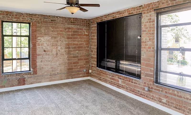 Empty room with exposed brick walls, three windows with black blinds, a ceiling fan with light fixture, and carpeted floor.