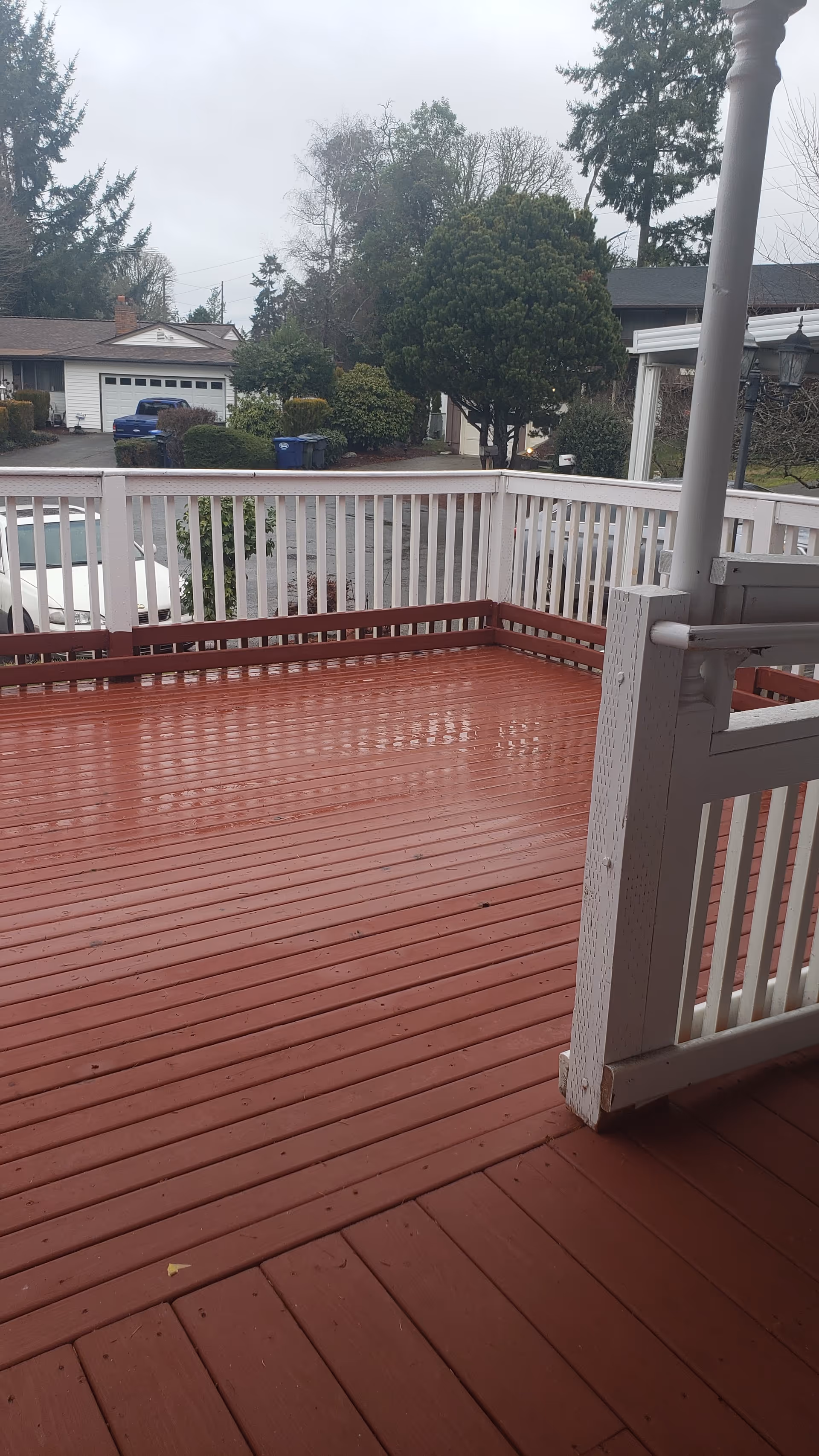 A wet wooden deck with red floorboards and white railing, overlooking a residential street with houses, trees, and parked cars on a cloudy day.