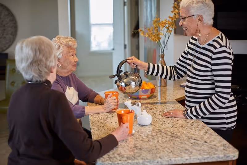 Three elderly women gathered around a kitchen island, one woman is pouring tea from a kettle into orange cups held by the other two women, with a bowl of oranges and a vase with yellow flowers on the countertop.