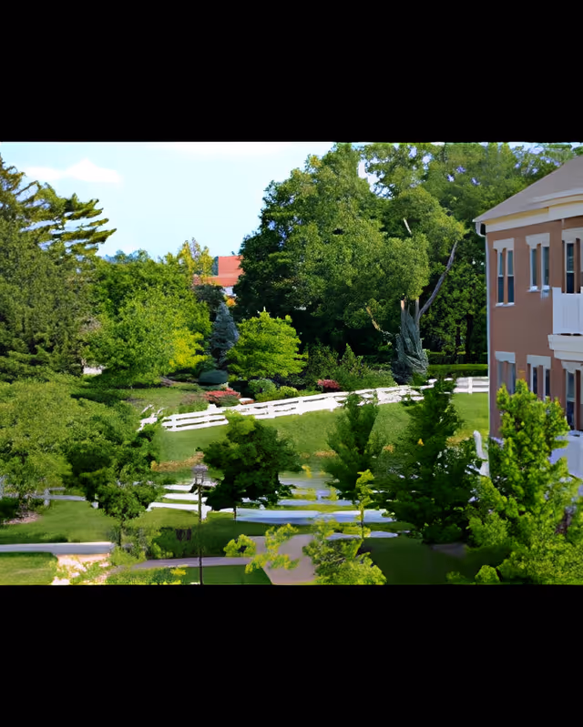 A scenic outdoor view of a senior living facility with lush green trees, manicured lawns, a white wooden fence, and a multi-story building with balconies on the right side.