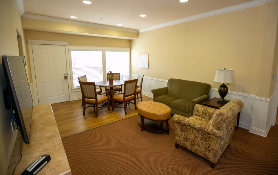 Small senior living common room with a sofa, armchair, ottoman, dining table and entry door.