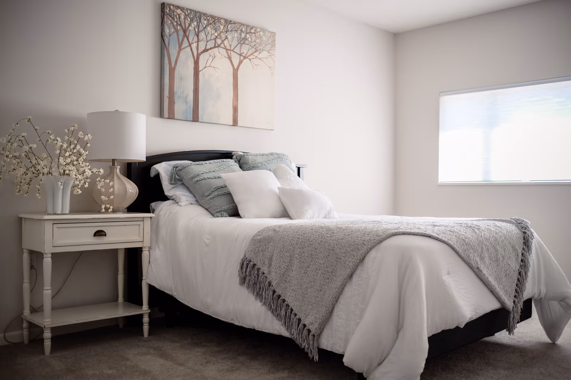 A neatly made bed with white bedding, a gray throw blanket, and several pillows in a bright bedroom. Next to the bed is a white nightstand with a beige lamp and a vase with white flowers. A painting of trees hangs on the wall above the bed, and a window with a closed blind lets in natural light.