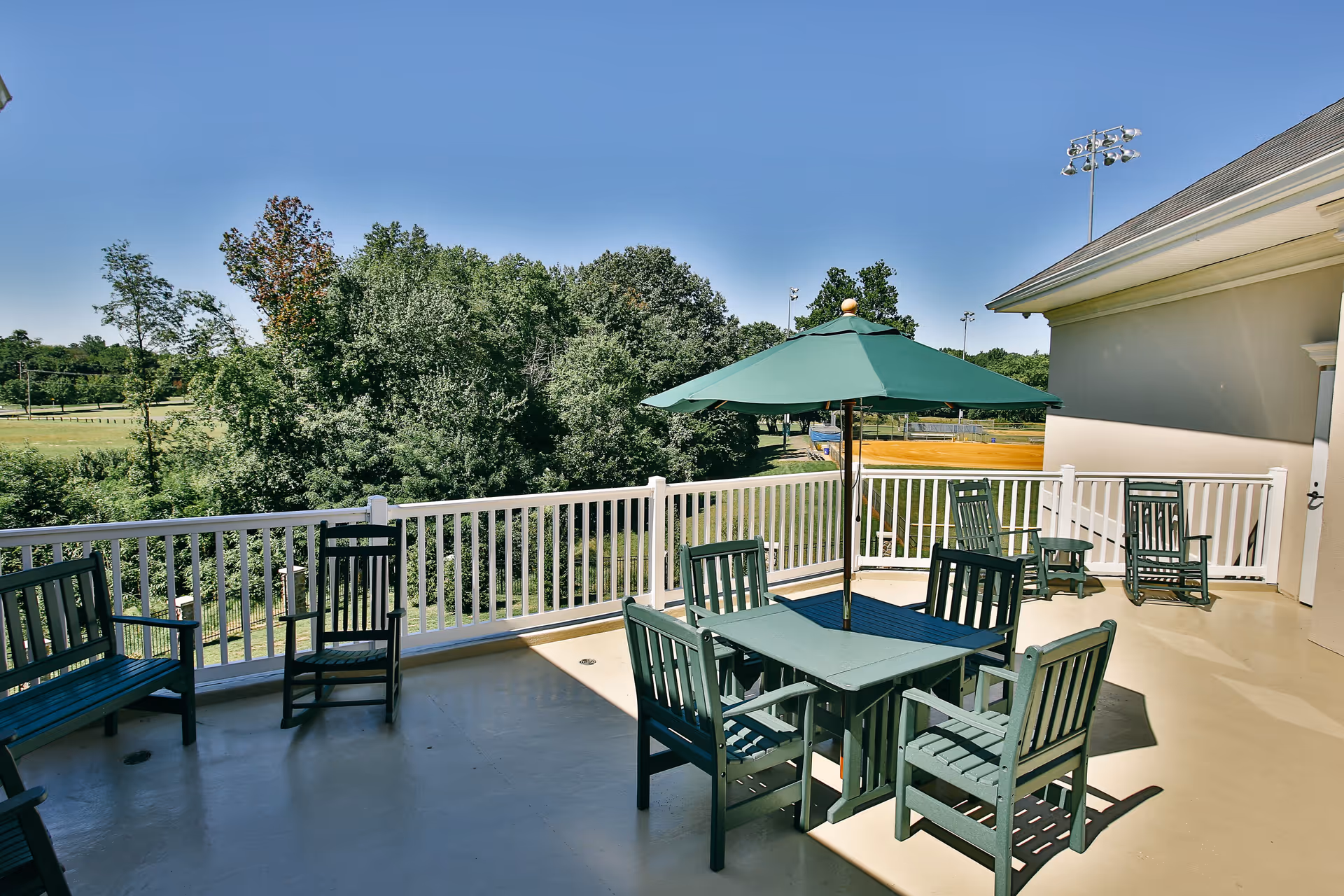 Outdoor balcony patio with green table, chairs, umbrella and white railing overlooking trees and sports fields.