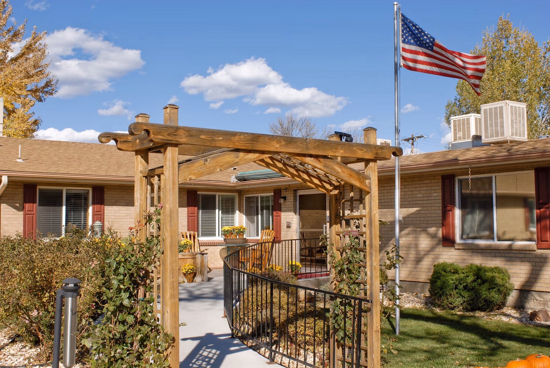 Outdoor view of Canon Lodge Care Center showing a wooden pergola over a walkway leading to the entrance. The building has beige brick walls with red shutters on the windows. An American flag is flying on a flagpole to the right, and there are bushes and plants along the walkway under a clear blue sky with some clouds.